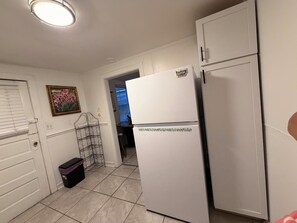 A kitchen with white cabinets, a white refrigerator, a black trash bin, and a decorative metal rack. - Sundance Tiny Home - Coastal Getaway by the Bay (Norfolk)