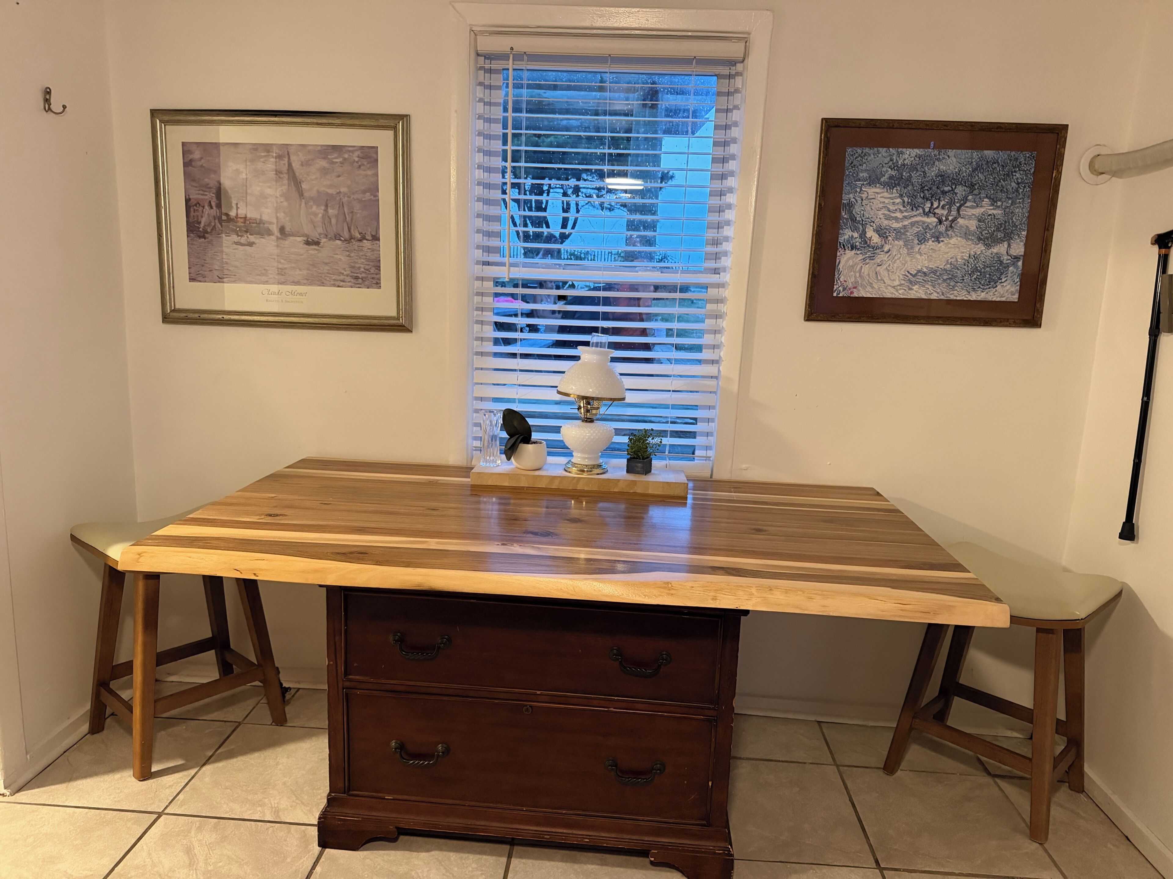 A wooden desk with drawers, a window with blinds, and two framed artworks on the wall.