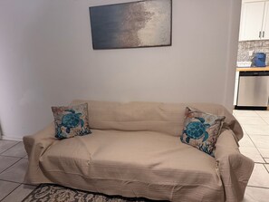 Un canapé beige agrémenté de coussins à motifs, un tableau abstrait accroché au mur et un coin cuisine équipé d’un lave-vaisselle.