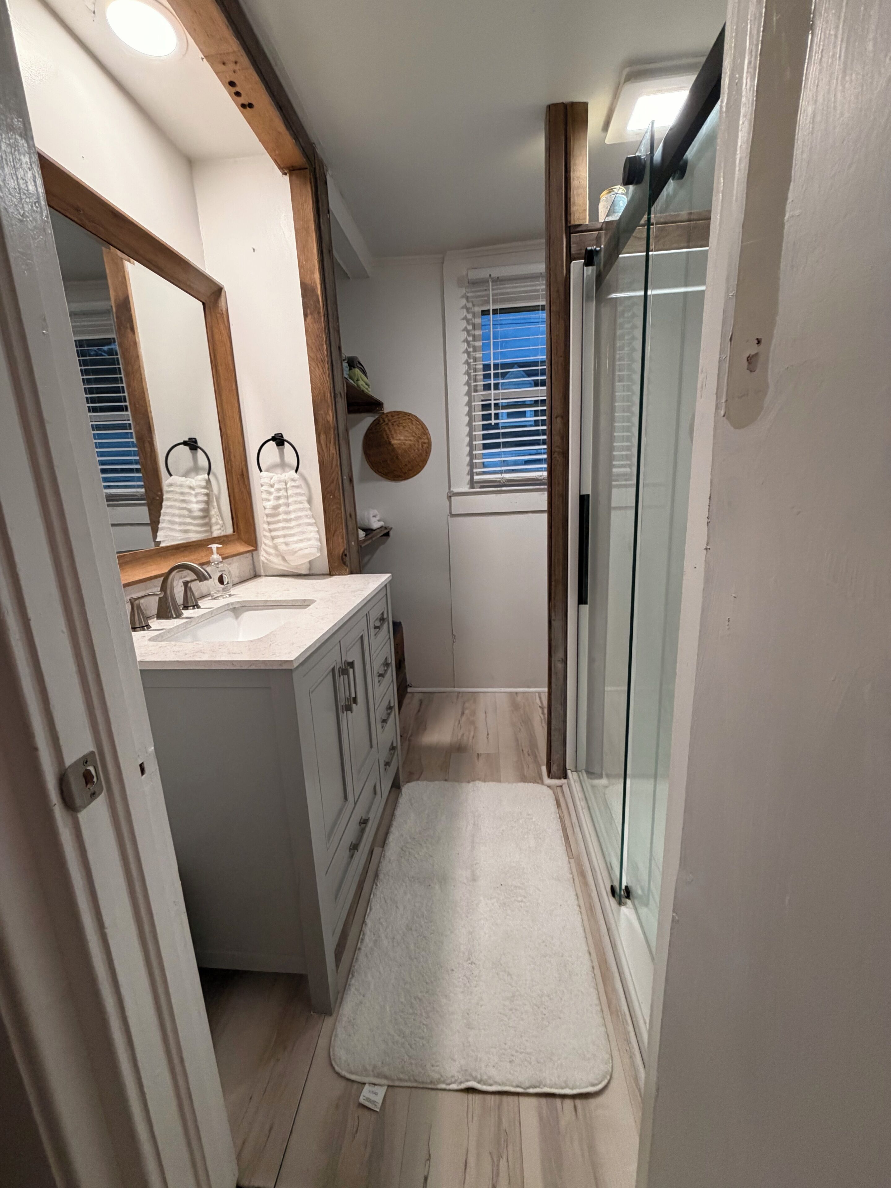 Un baño estrecho con lavamanos blanco, tocador de madera y cabina de ducha de vidrio.
