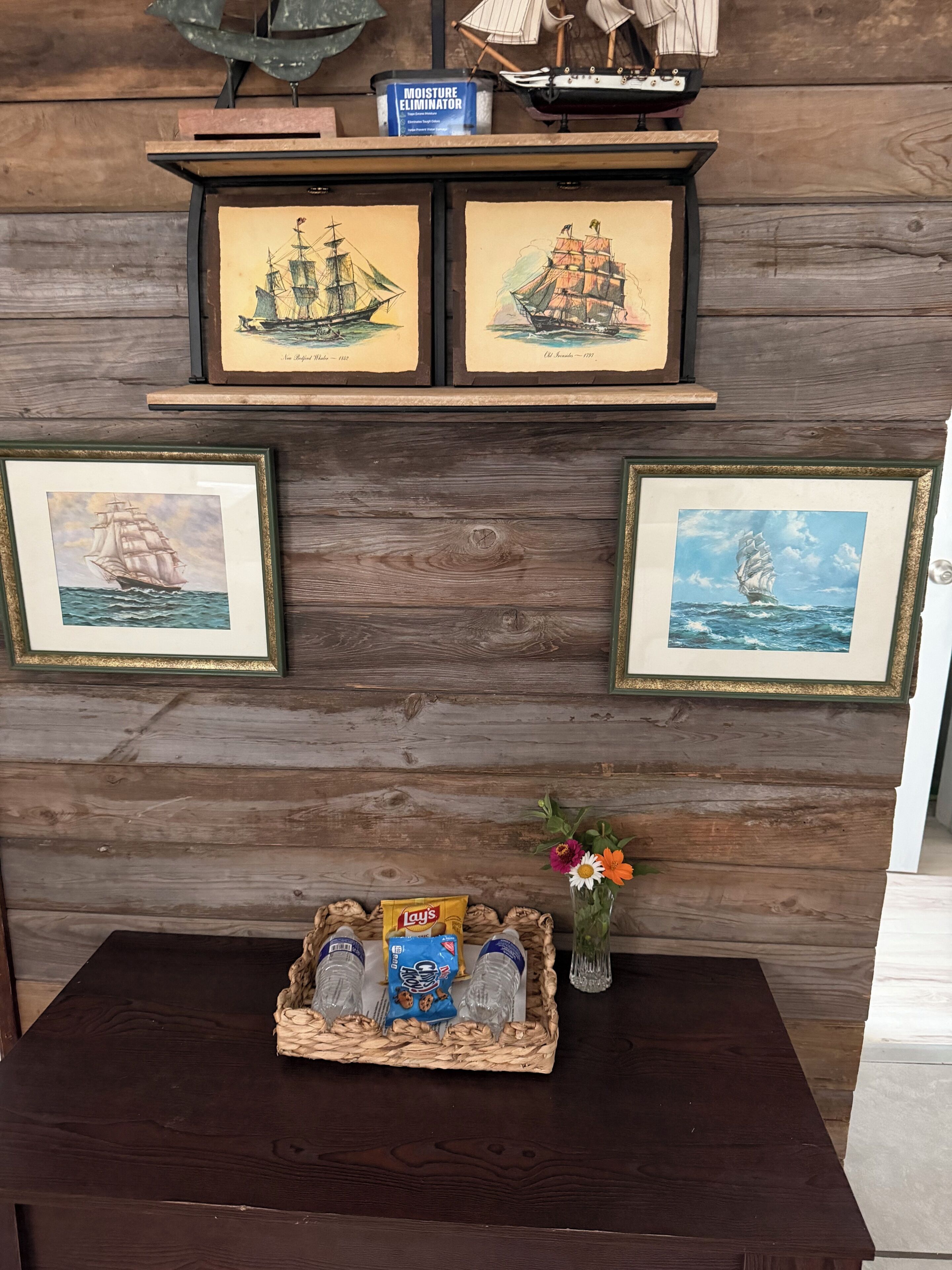 A wooden wall with framed maritime paintings and a small table with snacks and a vase of flowers.