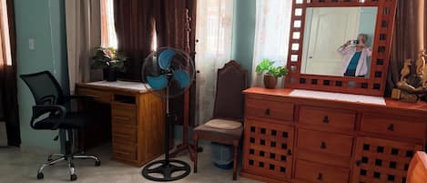 A room with a wooden desk, a chair, a standing fan, a dresser with a mirror, and a potted plant.