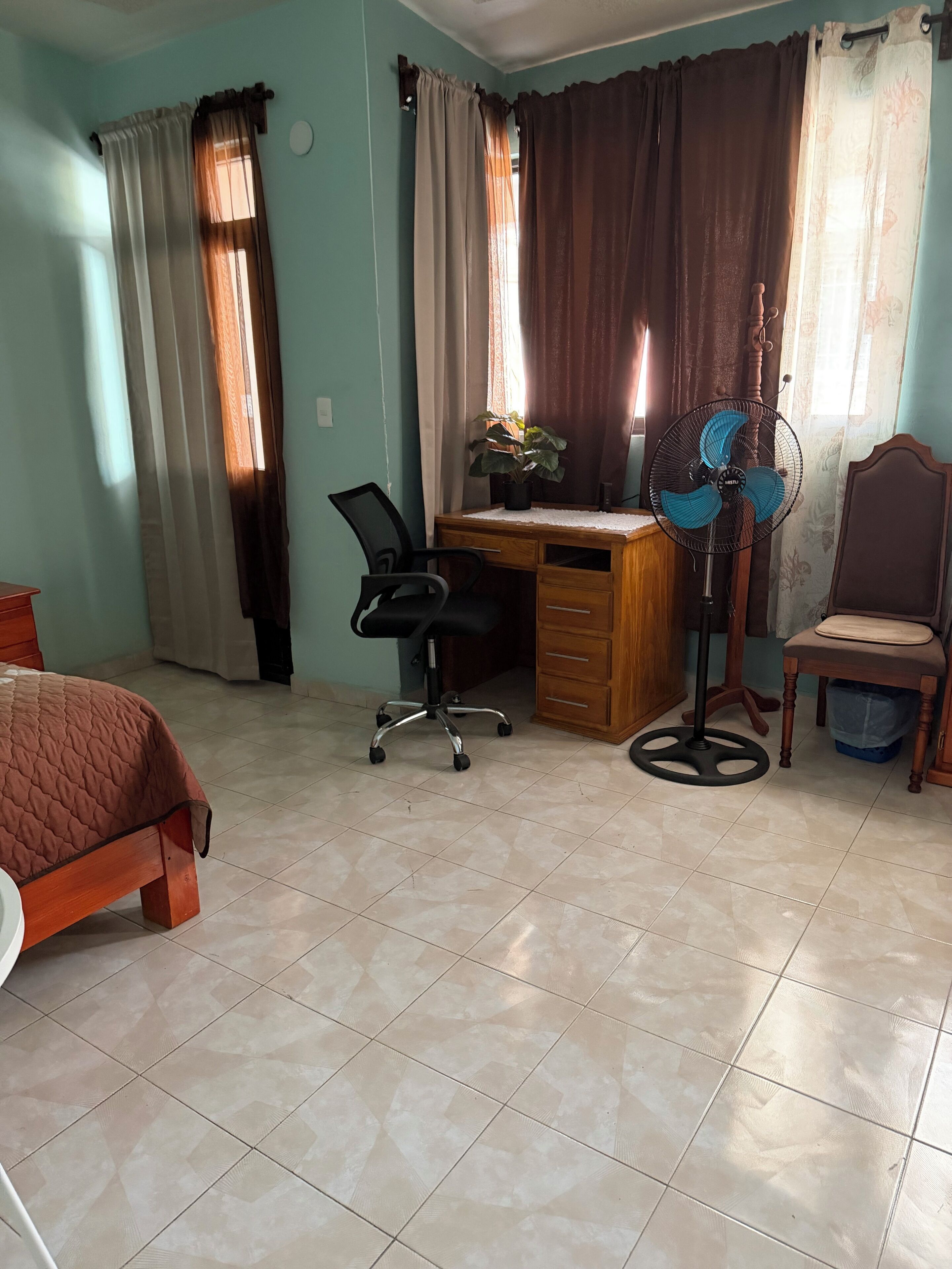 Une chambre à coucher comprenant un lit, un bureau, une chaise et un ventilateur sur pied.