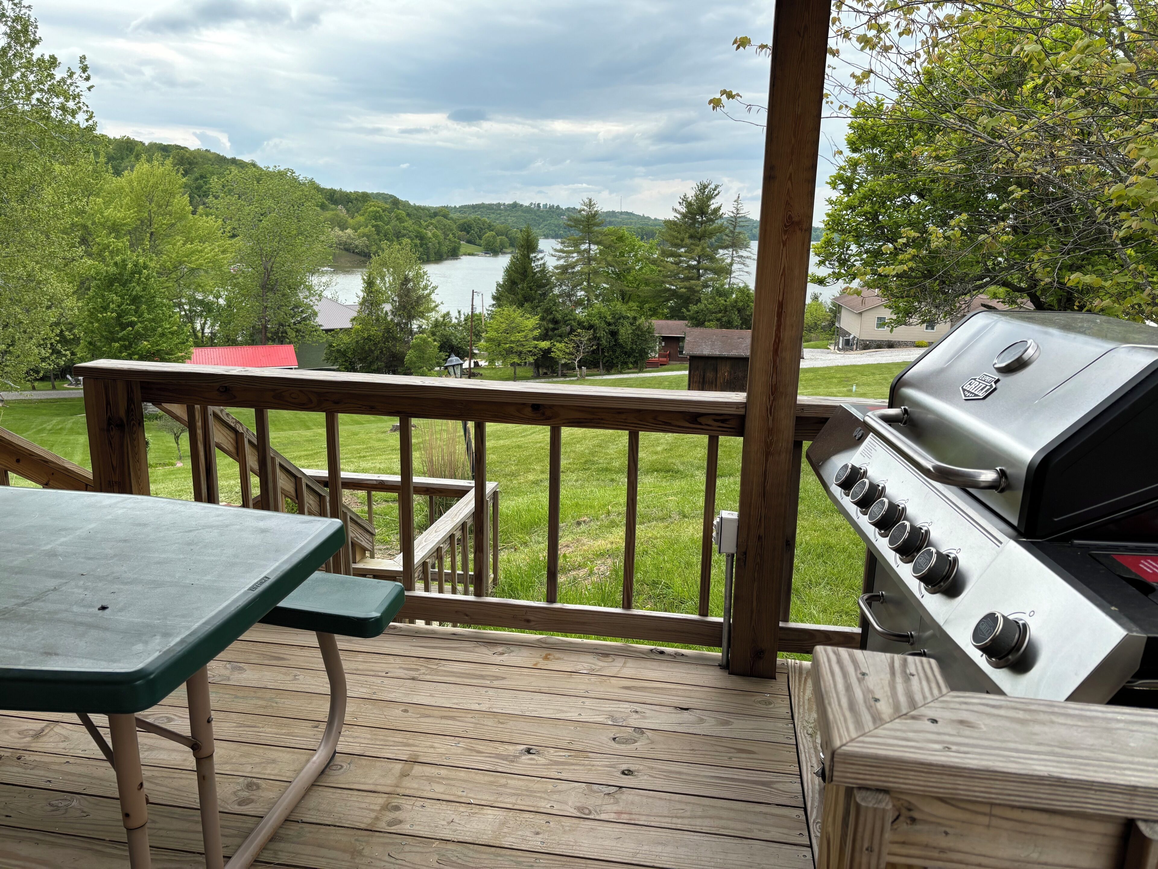 Eine Holzterrasse mit Grill und Tisch, mit Blick auf einen See und bewaldete Hügel.