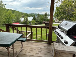A wooden deck with a grill and a table, overlooking a lake and forested hills.