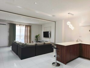 A modern living room with a sectional sofa, a TV, and a kitchen area with wooden cabinets and a bar stool. - Horizon view (Flic en Flac)