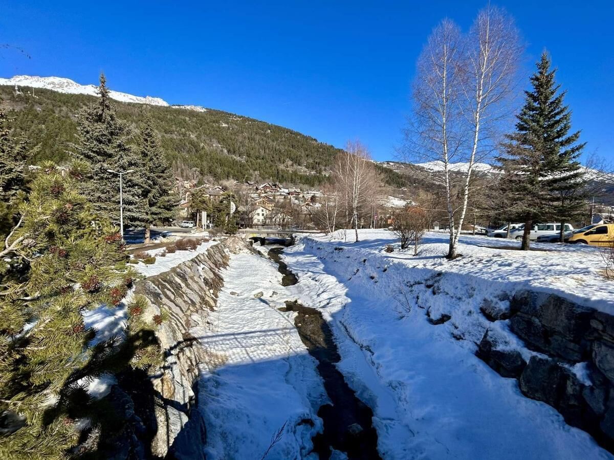 A snow-covered landscape with a stream, trees, and a mountain in the background.