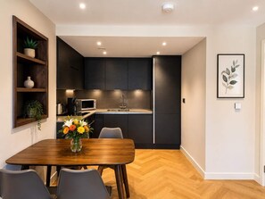 A modern kitchen with dark cabinetry, a microwave, and a sink. There is a dining table with chairs and a vase of flowers. - The Pullman Suite - near train station (York)