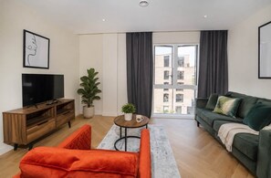 A modern living room with a flat-screen TV, a wooden entertainment unit, a green sofa, an orange armchair, and a small round table with a plant. - The Pullman Suite - near train station (York)