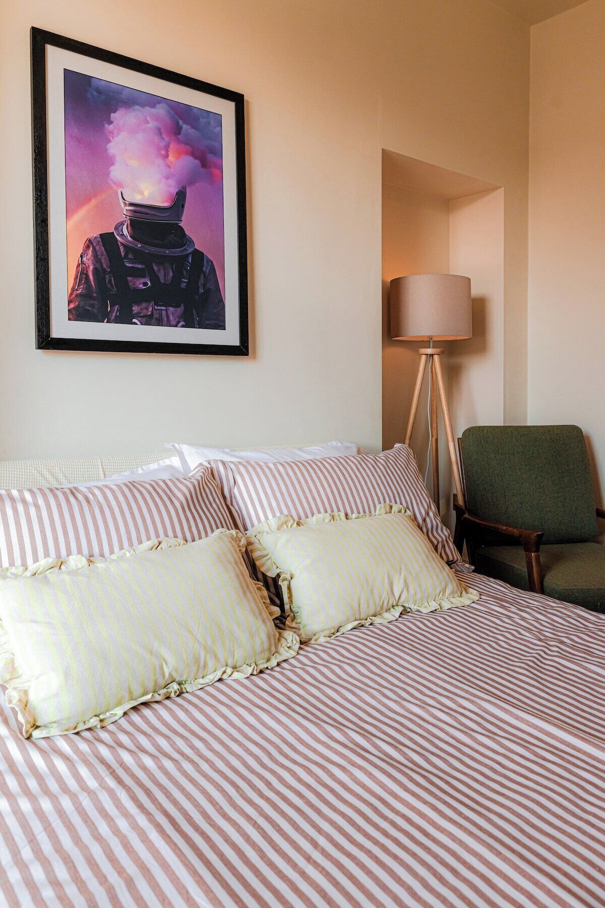 Hotelkamer met een bed, kussens, een lijstje aan de muur, een vloerlamp en een stoel.