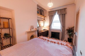 A bedroom with a bed, a shelf with plants, a fireplace, and a window with curtains.