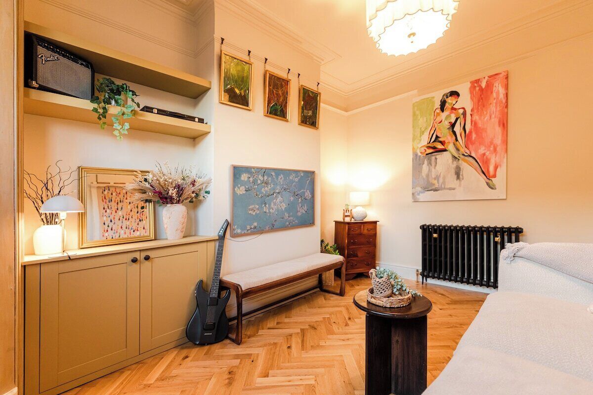 A room with a wooden herringbone floor, a bench, a guitar, a radiator, and a painting on the wall.