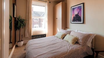Een slaapkamer met een bed, een raam, een radiator, een kamerplant en een ingelijst kunstwerk aan de muur.