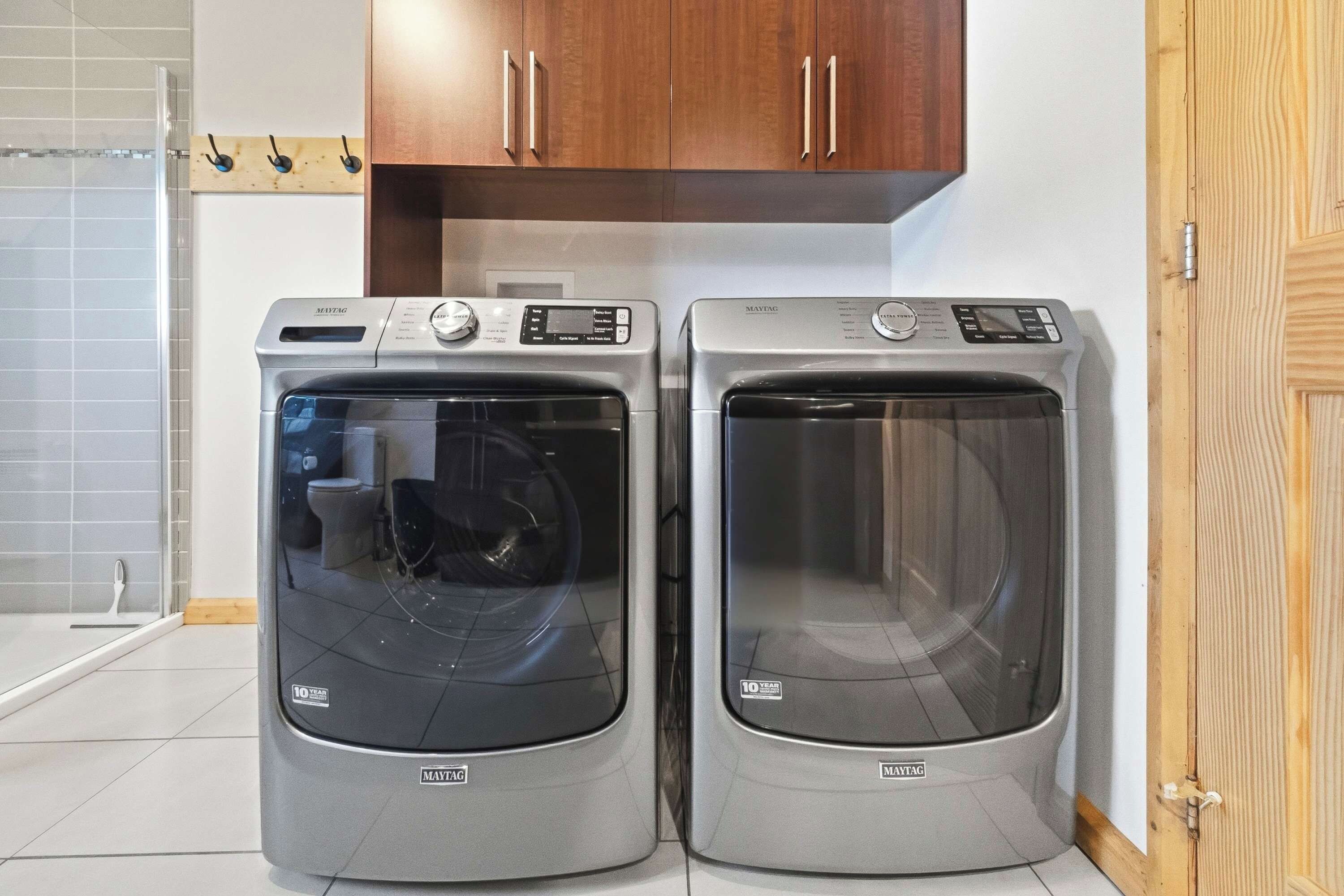 Two Maytag front-loading washing machines in a laundry room with a wooden cabinet above and a tiled wall.