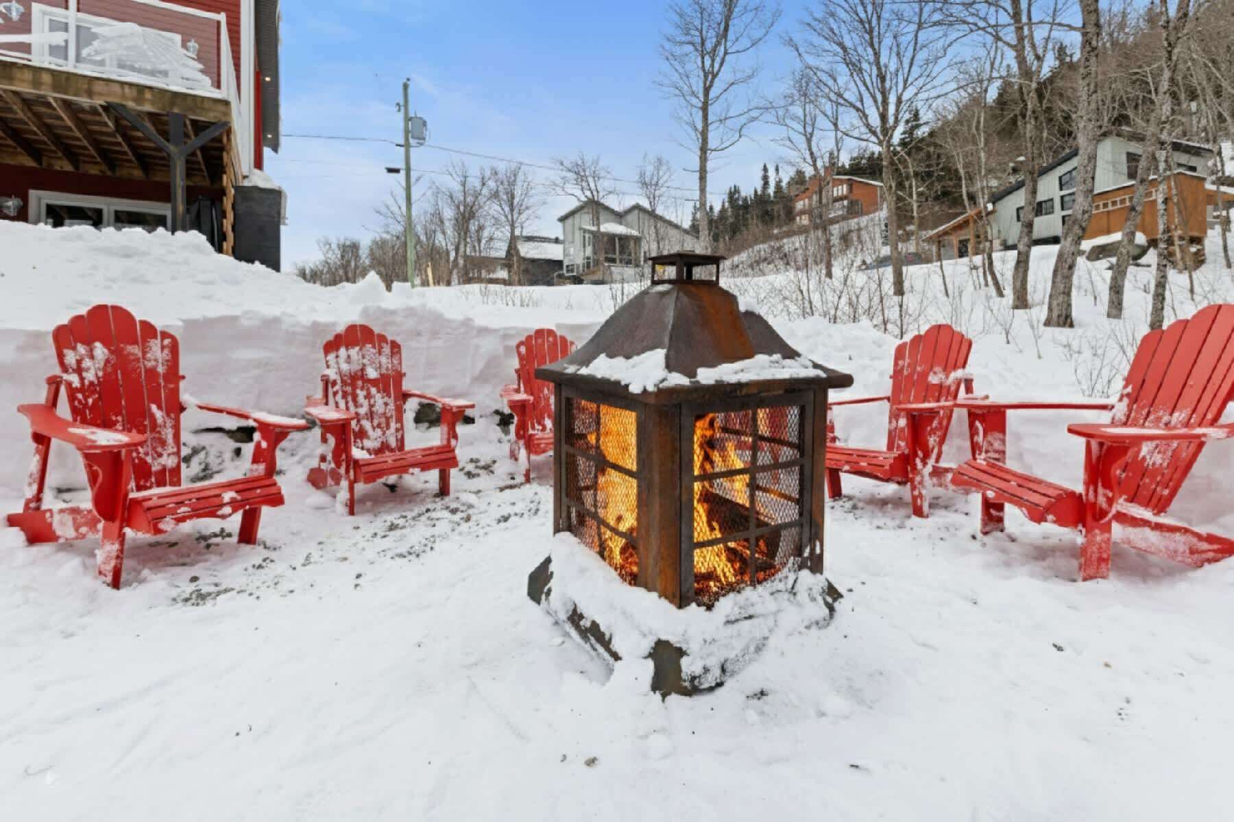 Un espace extérieur aménagé avec des sièges, recouvert de neige, et doté d’un foyer central entouré de chaises rouges.