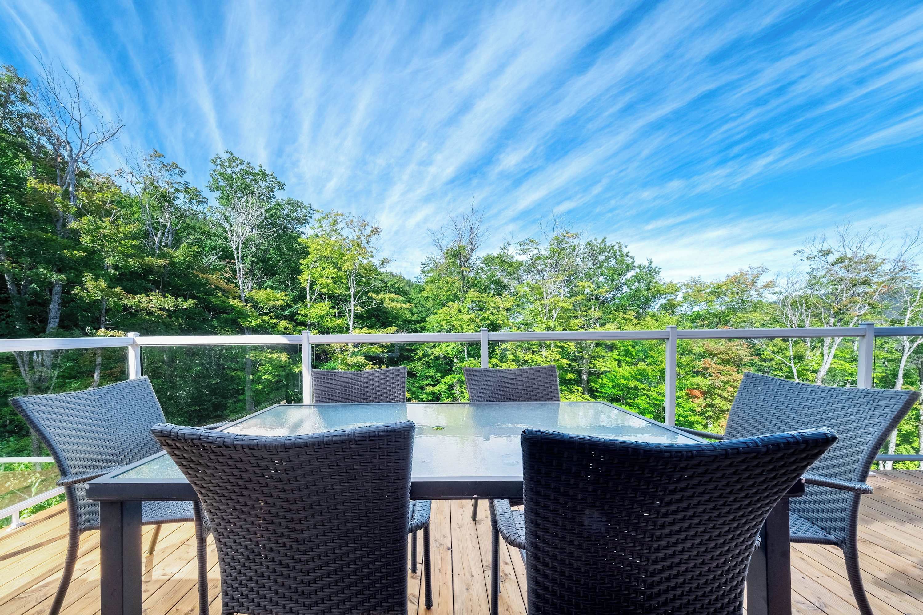 An outdoor deck with a glass-top table and wicker chairs, surrounded by a white railing and lush greenery.