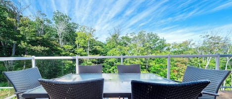 An outdoor deck with a glass-top table and wicker chairs, surrounded by a white railing and lush greenery.