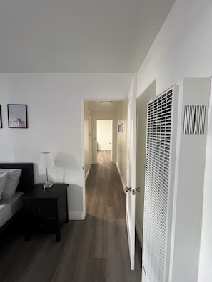 A hotel room with a bed, bedside table, lamp, and a view of a hallway leading to a door.