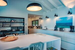 A modern kitchen with a dining area, featuring a white table, blue cabinets, and a large window with a view of the ocean. - Villa Summertime (2 bedrooms) (Saint Barthélemy)