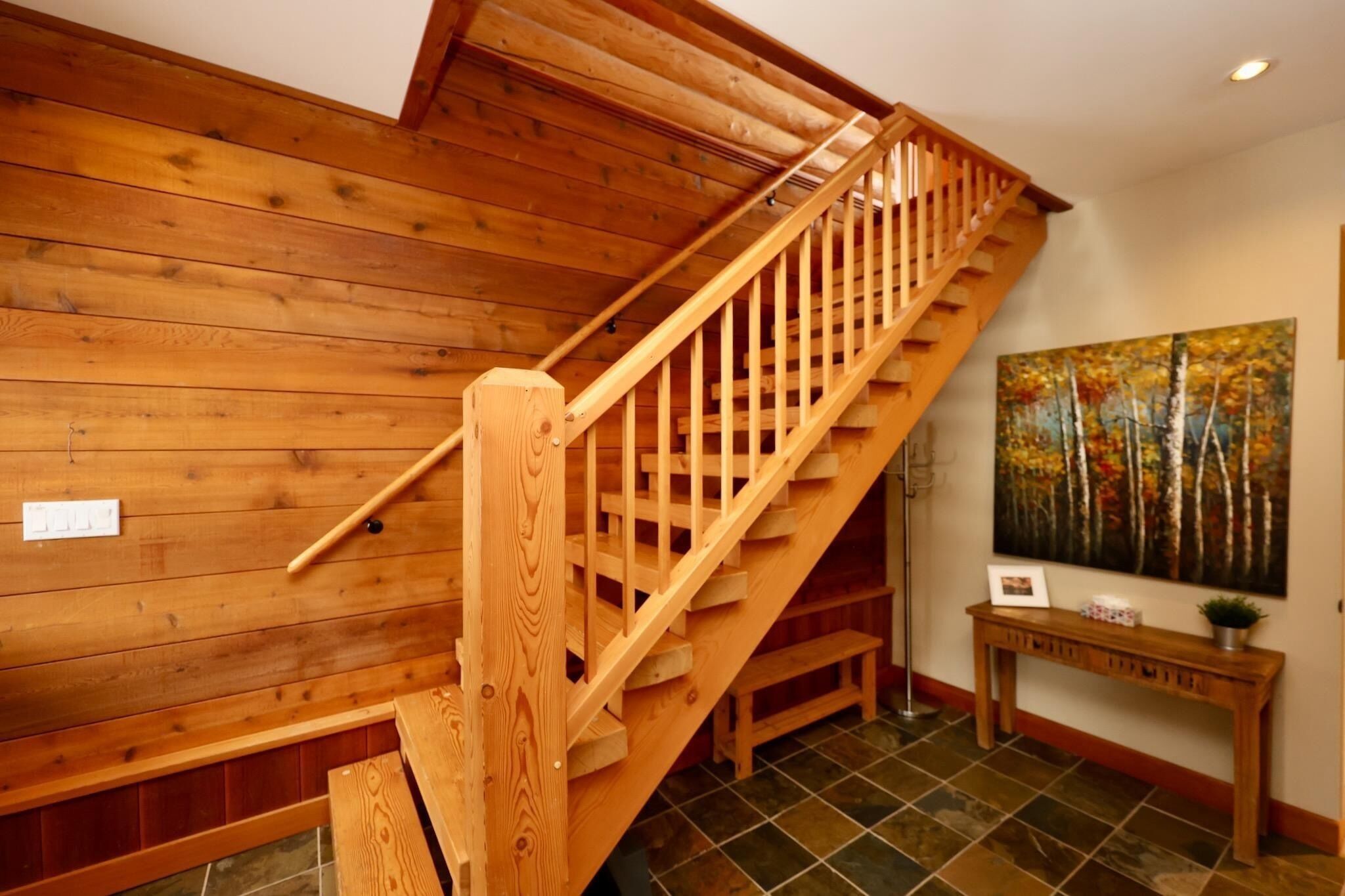 A wooden staircase with a handrail, a wooden wall paneling, a tiled floor, a painting of trees, and a small wooden console table with a plant.