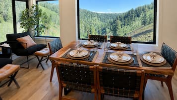 Un comedor con una mesa dispuesta para cuatro, ventanas amplias con vistas a un bosque, y una zona de estar con una silla negra y una planta.