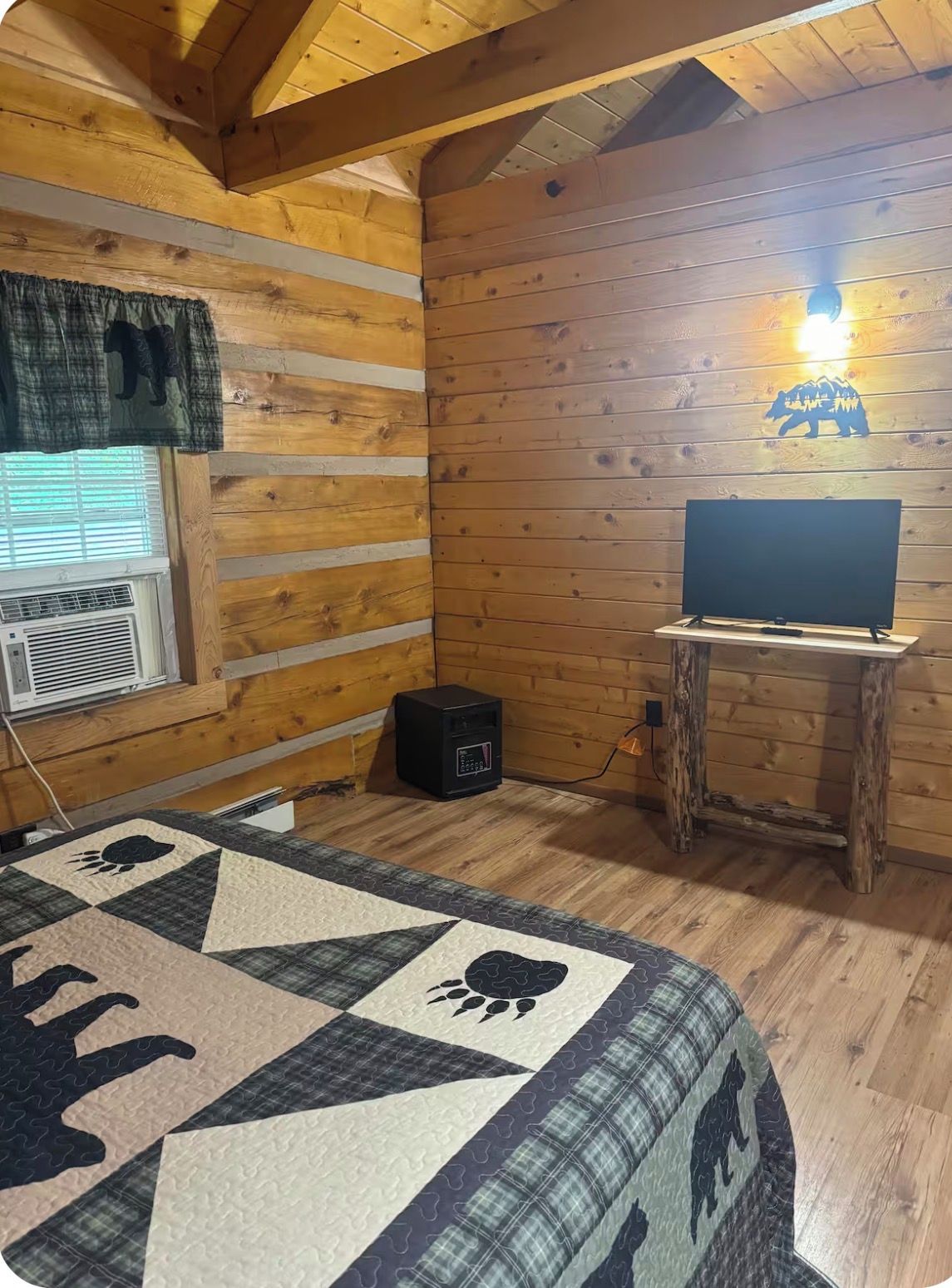 Ein Zimmer einer Holzhütte mit gesteppter Bettdecke, einem Fernseher auf einem hölzernen Standfuß und einer wandmontierten Leuchte.