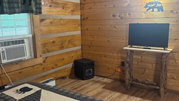 Ein Zimmer einer Holzhütte mit gesteppter Bettdecke, einem Fernseher auf einem hölzernen Standfuß und einer wandmontierten Leuchte.