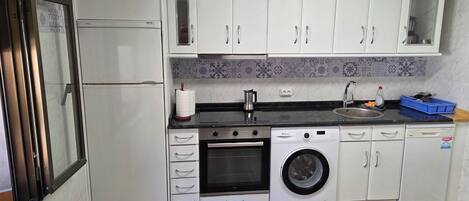 Cocina compacta con armarios blancos, horno integrado, lavadora y un lavabo.
