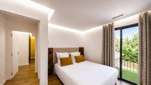 Une chambre avec un grand lit, un plancher en bois, une porte donnant sur une autre pièce et une vue sur la verdure à l’extérieur.