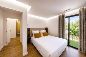 A bedroom with a large bed, a wooden floor, a door leading to another room, and a view of greenery outside. - La Bâtisse - Piscine & Jardin - Luberon (Gordes)