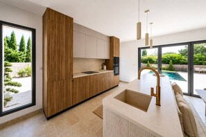 A modern kitchen with wooden cabinets, a built-in oven, and a large window overlooking a garden. - La Bâtisse - Piscine et jardin - Luberon (Gordes)