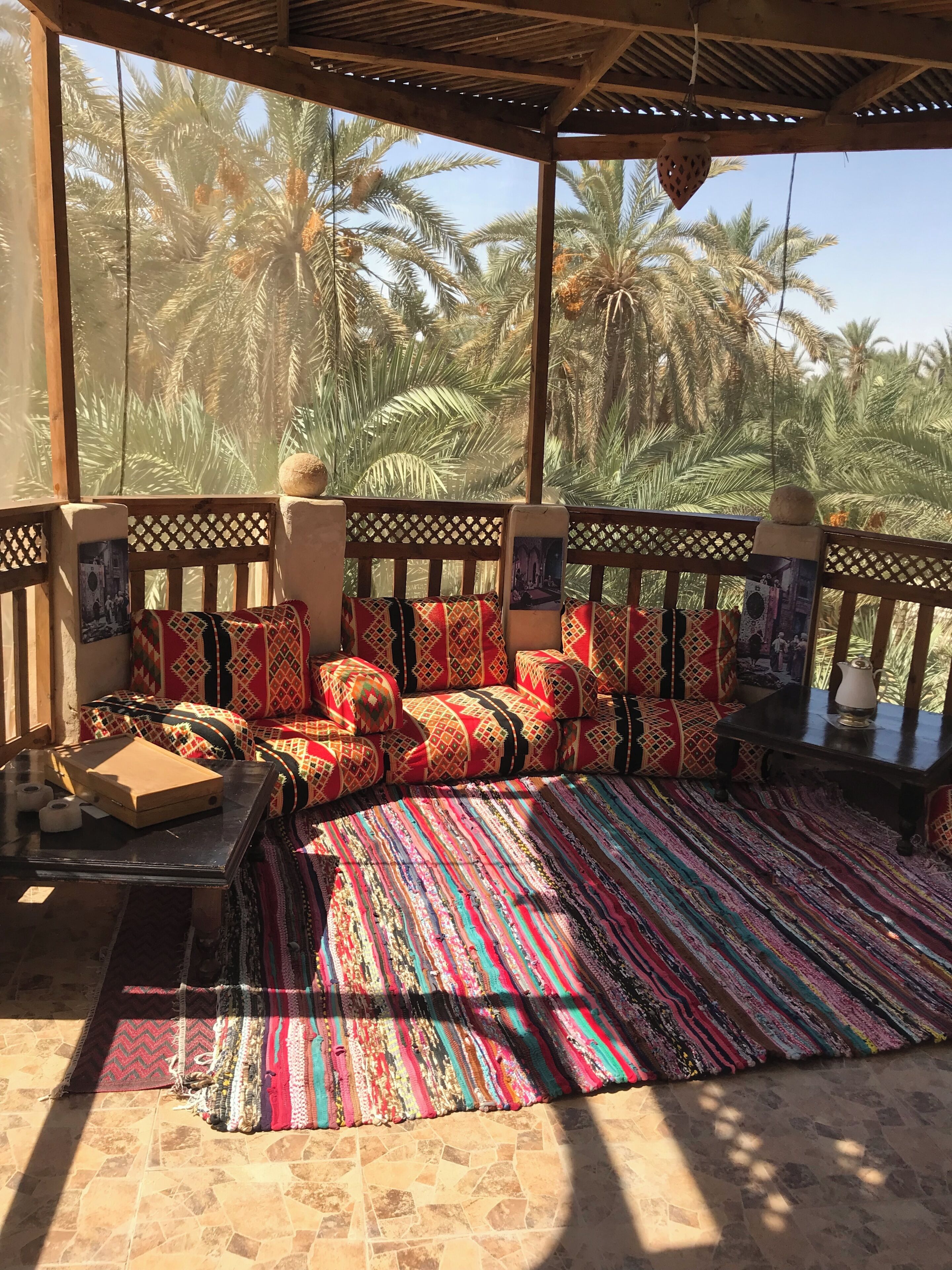 Un espacio de descanso techado con alfombra estampada, mobiliario de madera y vistas a palmeras.