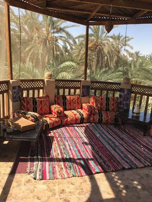 A covered seating area with a patterned rug, wooden furniture, and a view of palm trees.
