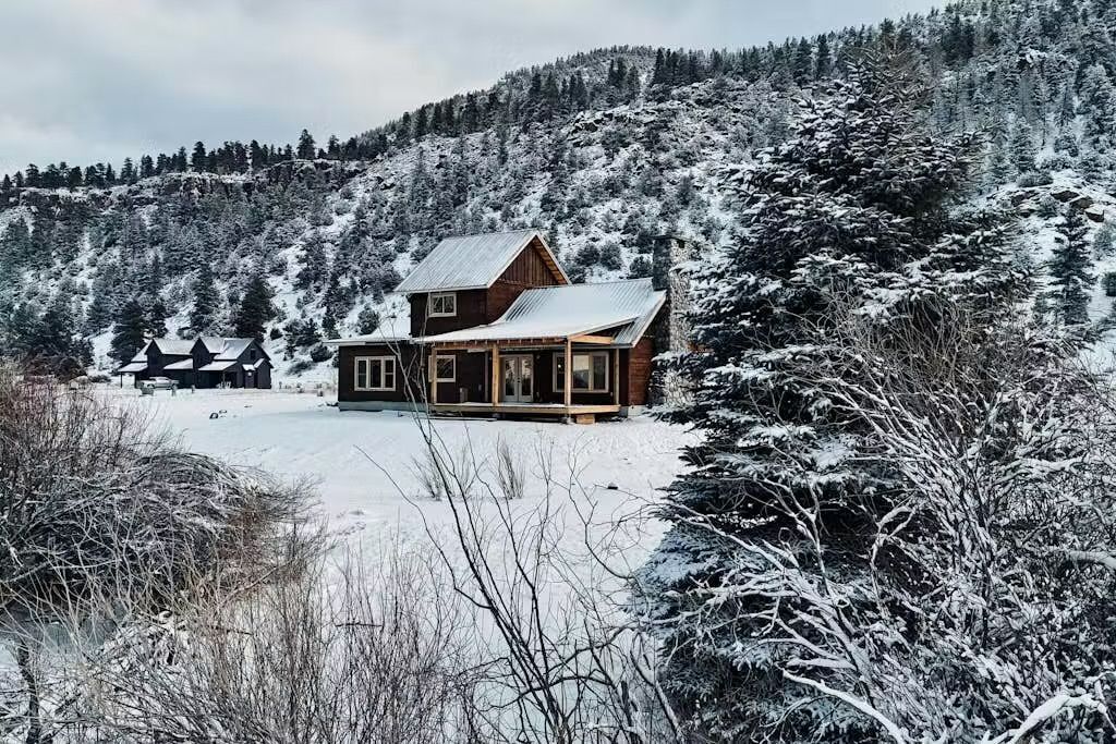 A wooden cabin with a metal roof, surrounded by snow-covered trees and a hill in the background.