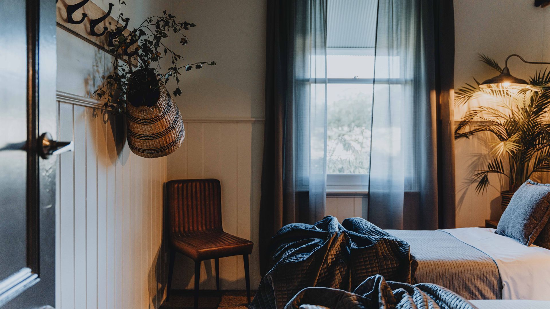Une chambre à coucher avec un lit, une chaise, une plante suspendue et une fenêtre avec des rideaux.