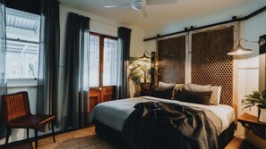 Un dormitorio con cama, una silla, ventana con cortinas, ventilador de techo y una planta.