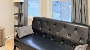 Une pièce avec un canapé en cuir noir, un grand lampadaire et une vue sur un arbre par la fenêtre.