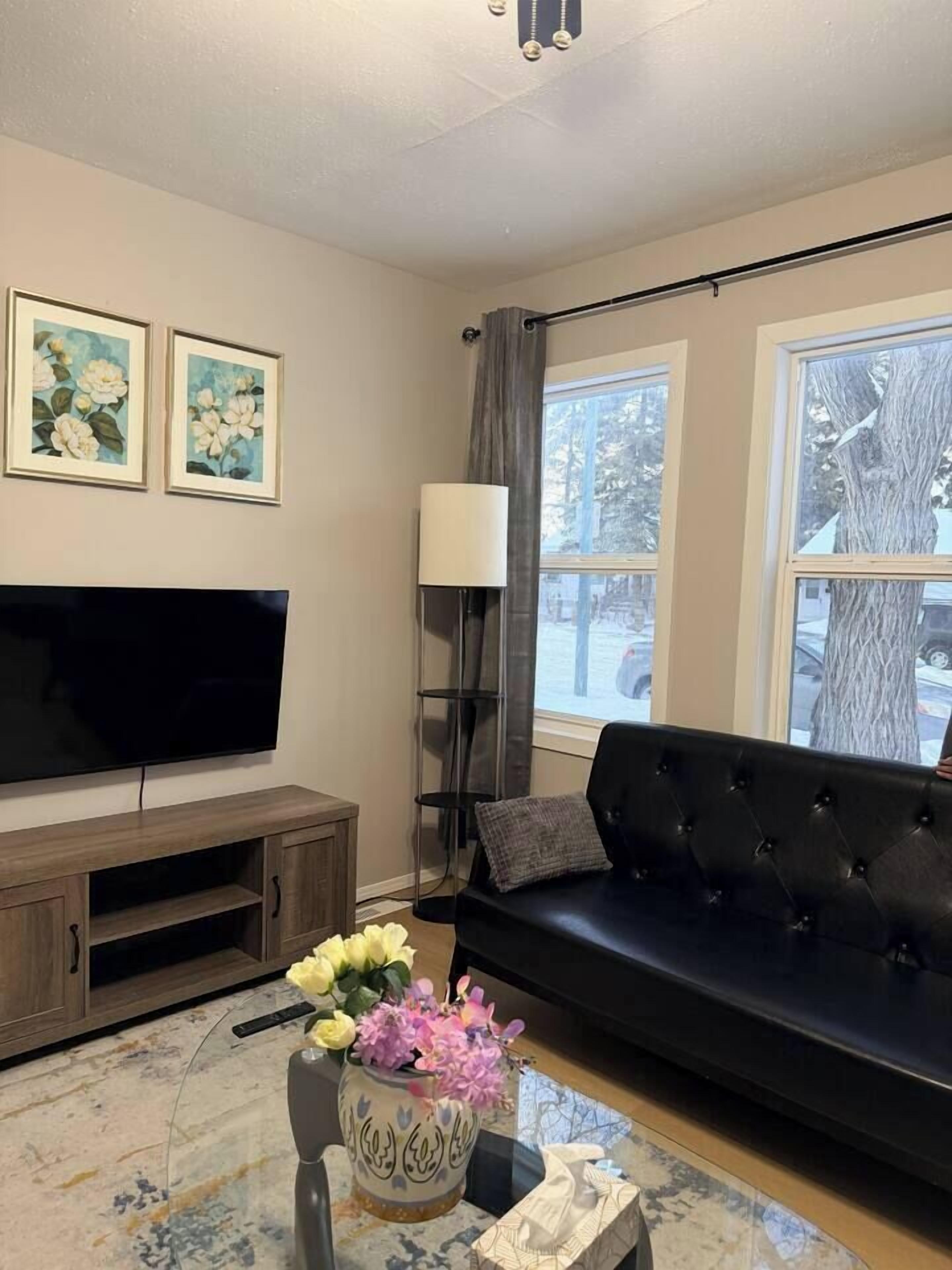 A living room with a flat-screen TV, a black sofa, a coffee table with a vase of flowers, and two framed floral paintings on the wall.