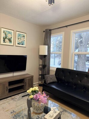 A living room with a flat-screen TV, a black sofa, a coffee table with a vase of flowers, and two framed floral paintings on the wall.