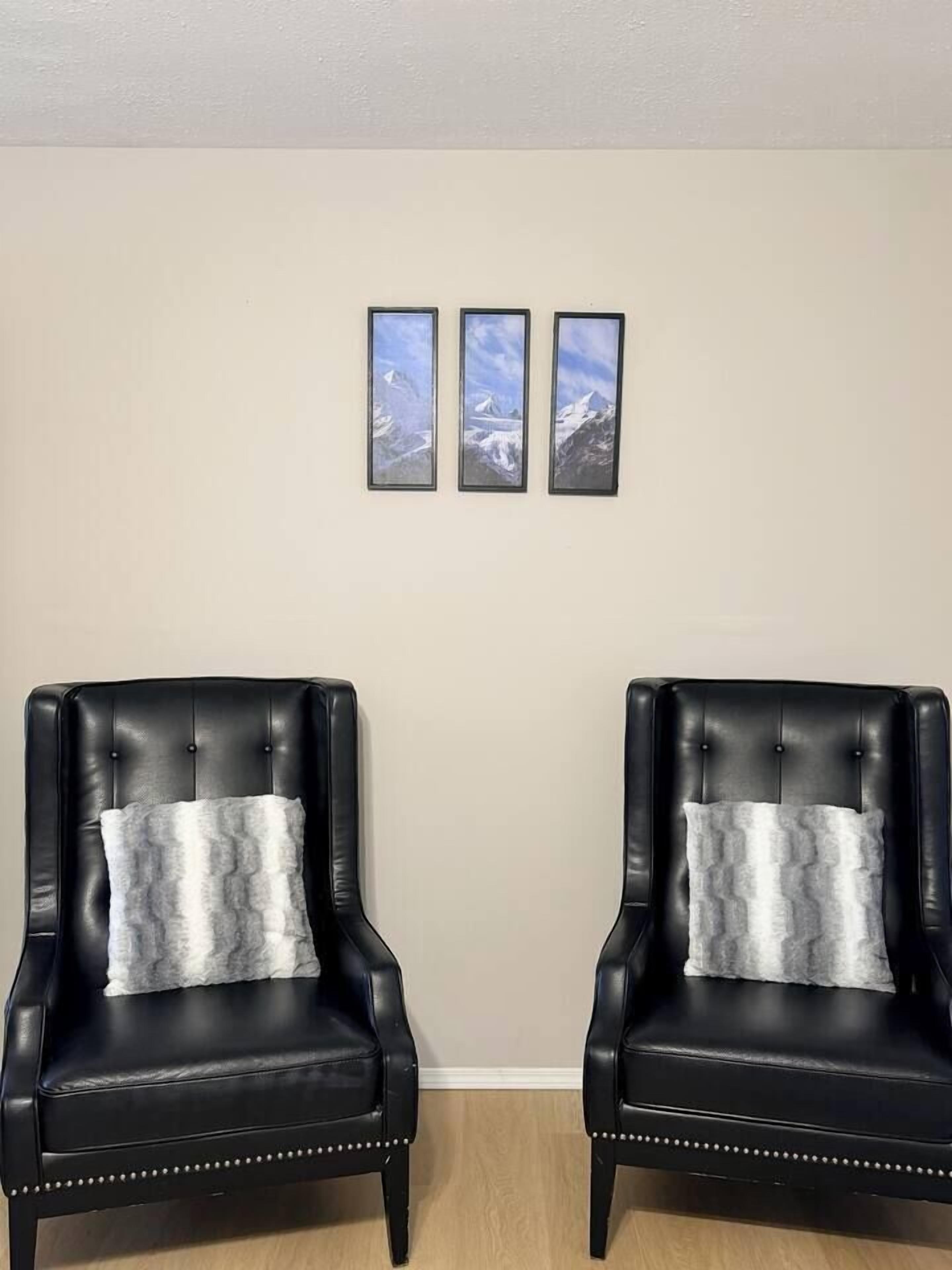 Deux fauteuils en cuir noir, dotés de coussins blancs, sont placés face à face, avec un mur beige en arrière-plan.