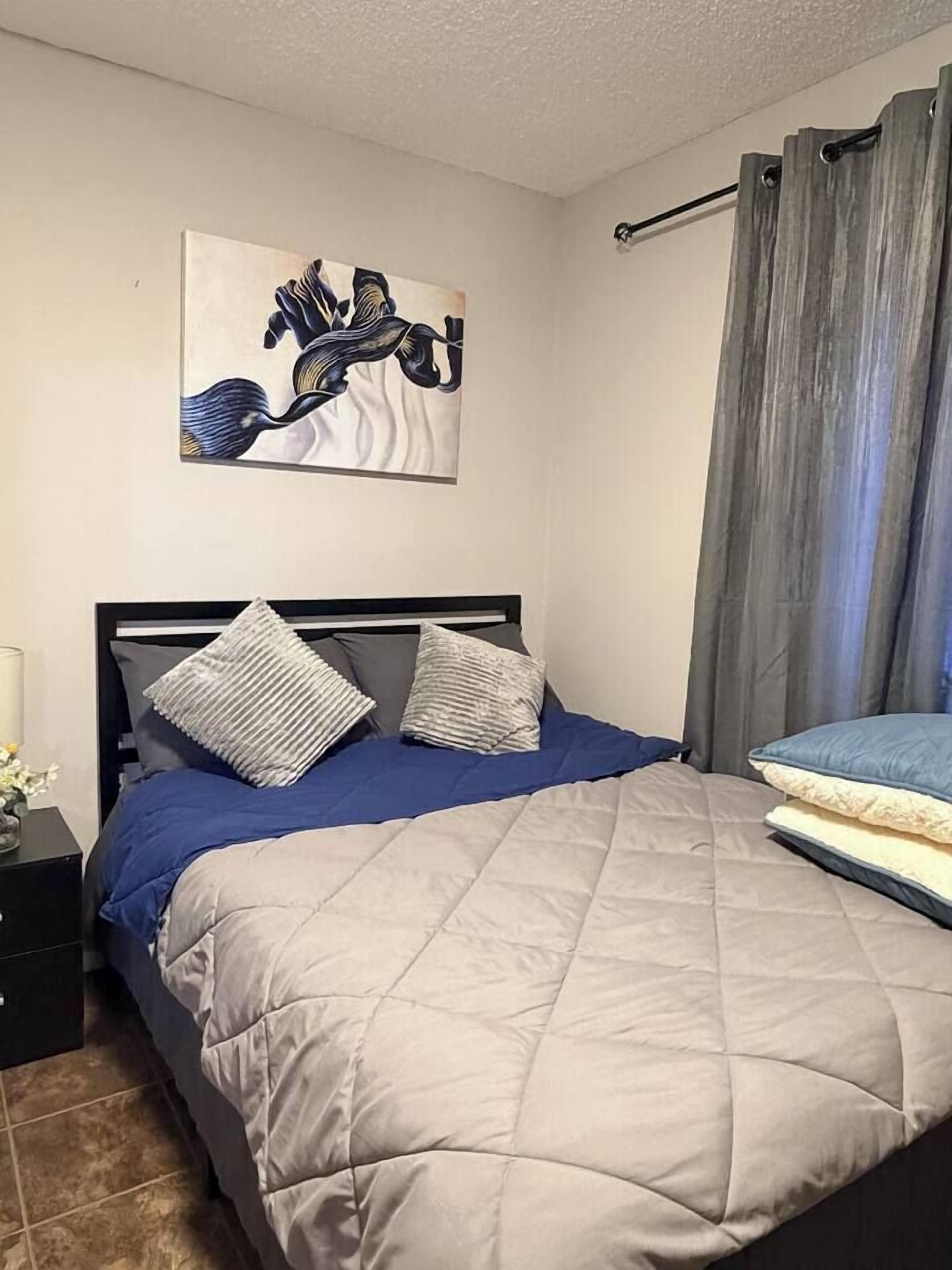 Un lit avec une couverture matelassée, deux oreillers, une tête de lit, une table de chevet avec une lampe et un tableau accroché au mur.