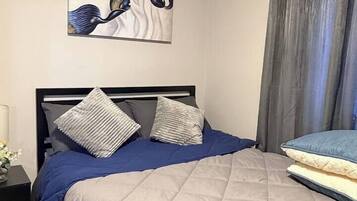 Un lit avec une couverture matelassée, deux oreillers, une tête de lit, une table de chevet avec une lampe et un tableau accroché au mur.