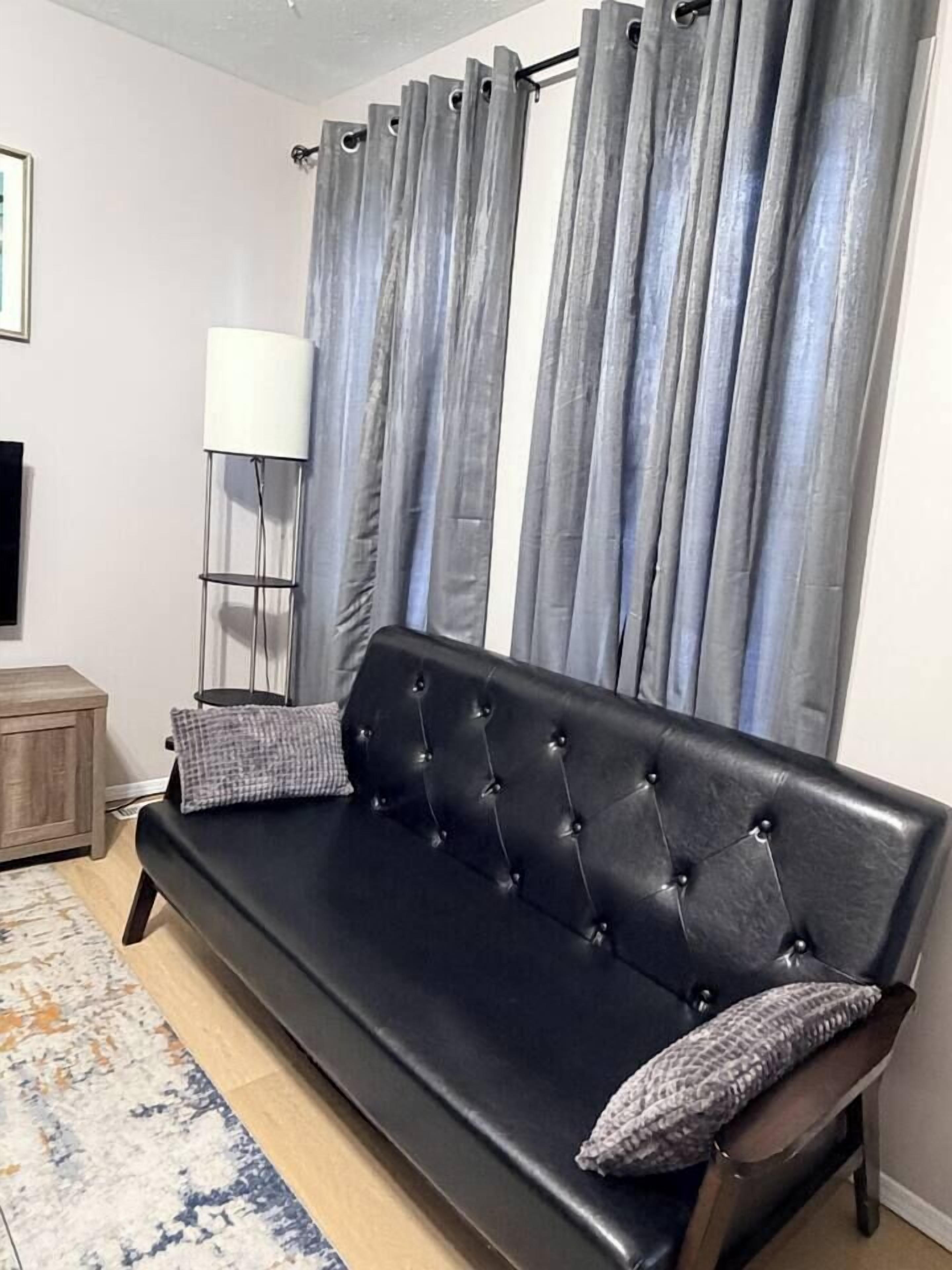 Un sofá negro con respaldo acolchado y dos almohadas, frente a una cortina gris.