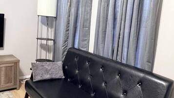 Un sofá negro con respaldo acolchado y dos almohadas, frente a una cortina gris.