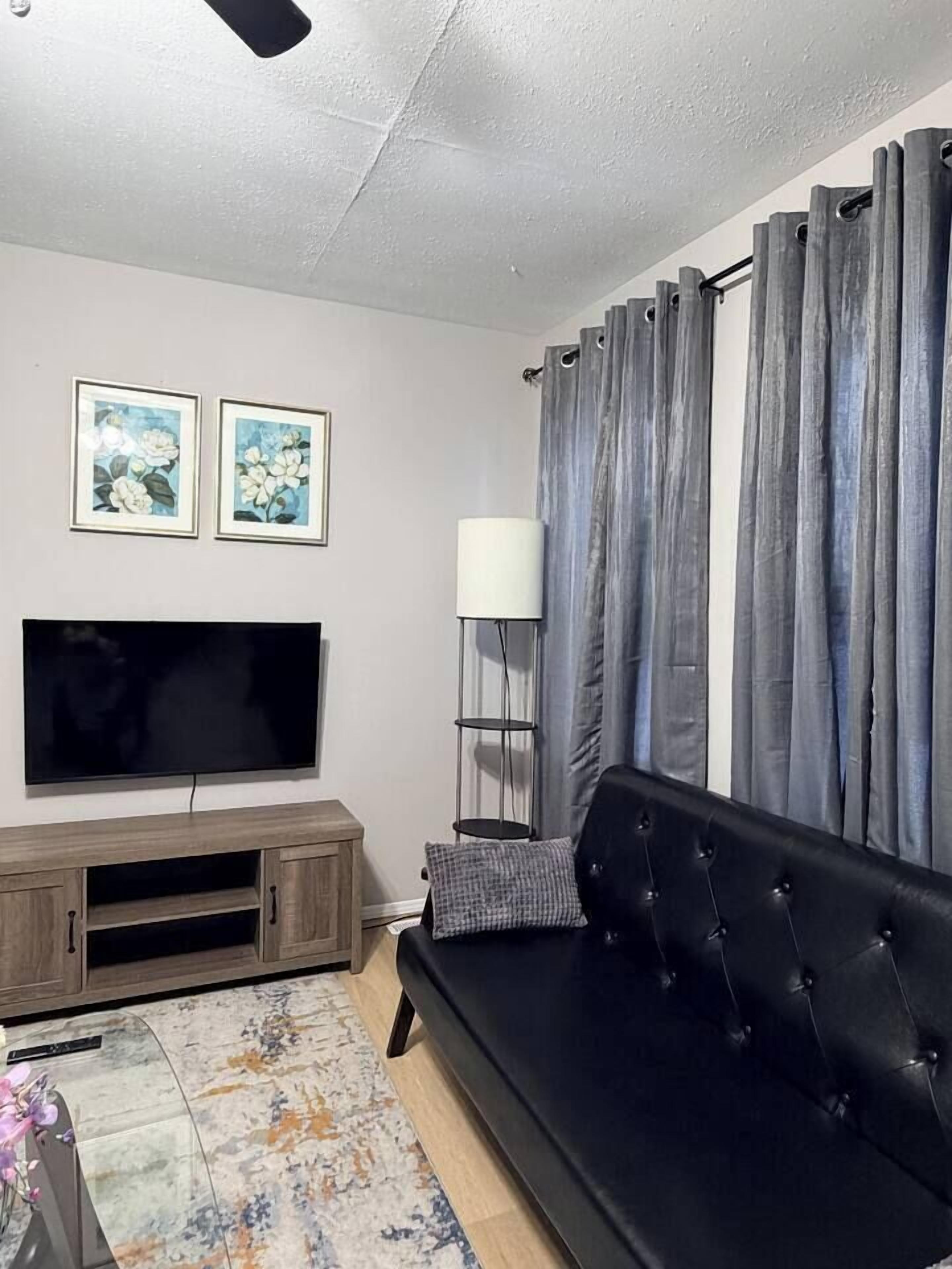 A living room with a flat-screen TV, a black sofa, grey curtains, and a floor lamp.