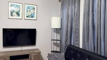 Un salon doté d’un téléviseur à écran plat, d’un canapé noir, de rideaux gris et d’un lampadaire.