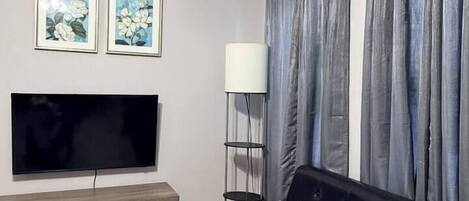 A living room with a flat-screen TV, a black sofa, grey curtains, and a floor lamp.
