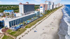 Ein Strandhotel mit mehreren Etagen, einem Poolbereich und einem Strand mit Liegestühlen.