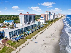 A beachfront hotel with multiple floors, a pool area, and a beach with sun loungers. - The Peachy Palm Oceanfront at Compass Cove (Myrtle Beach)