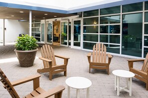 A modern outdoor seating area with wooden chairs and tables, a large potted plant, and a glass building facade. - The Peachy Palm Oceanfront at Compass Cove (Myrtle Beach)